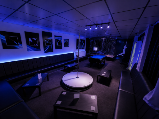 A dimly lit room with blue lighting, a pole, seating, and black boxes.